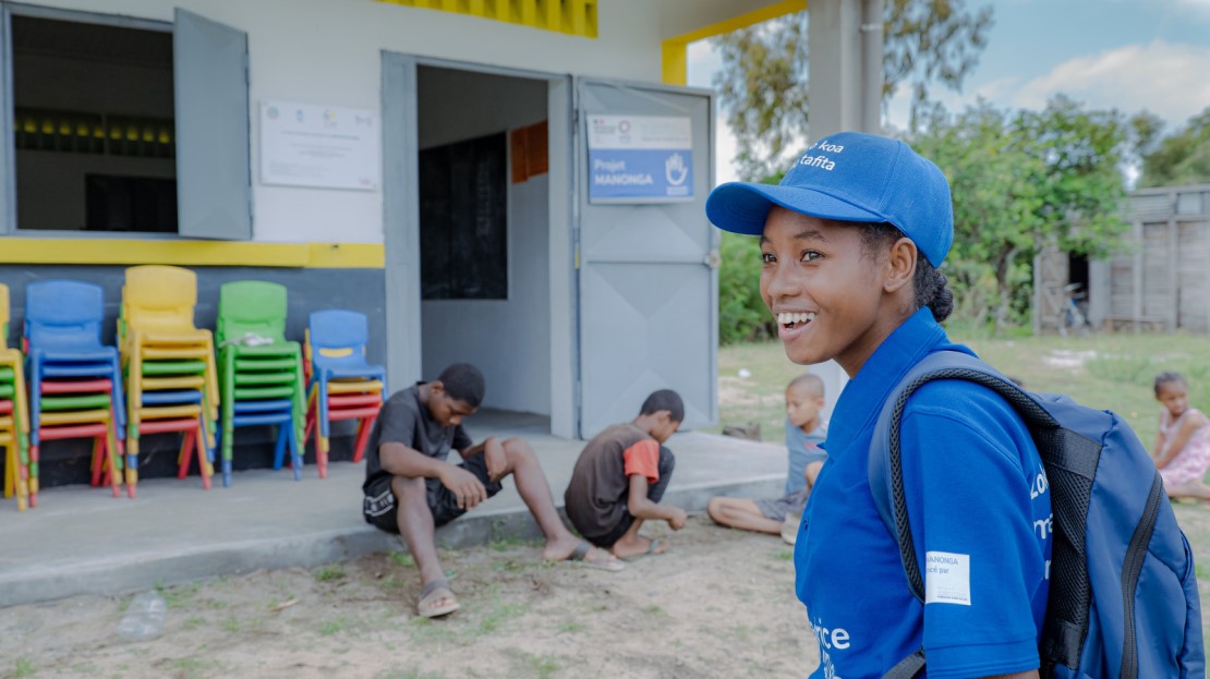 À Madagascar, une adolescente se tient debout devant son école et sourit. Des enfants sont assis sur le parvis.