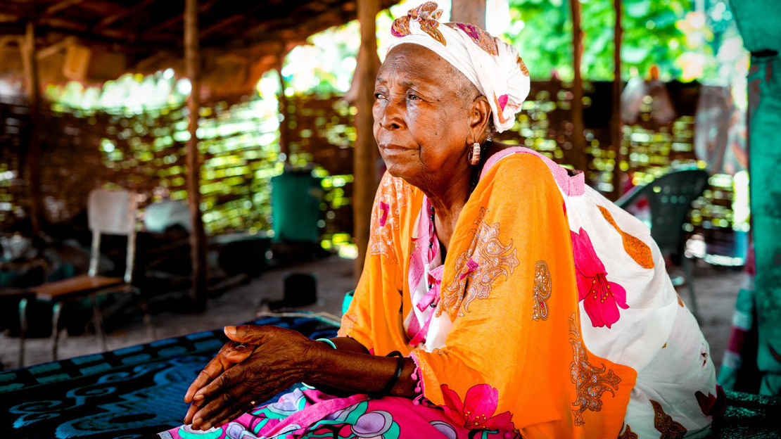 Portrait d'une femme âgée habillée de robe et de foulard colorés, assise à l'ombre d'une paillotte. Elle regarde vers la gauche et tient ses mains dans son giron.