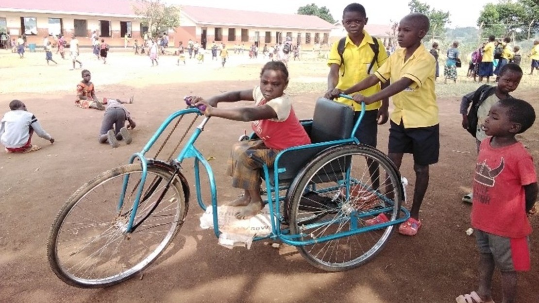 Une jeune fille est assise sur un tricycle bleu dans une cour d'école. Derrière elle, deux garçon en uniforme scolaire poussent le tricycle.