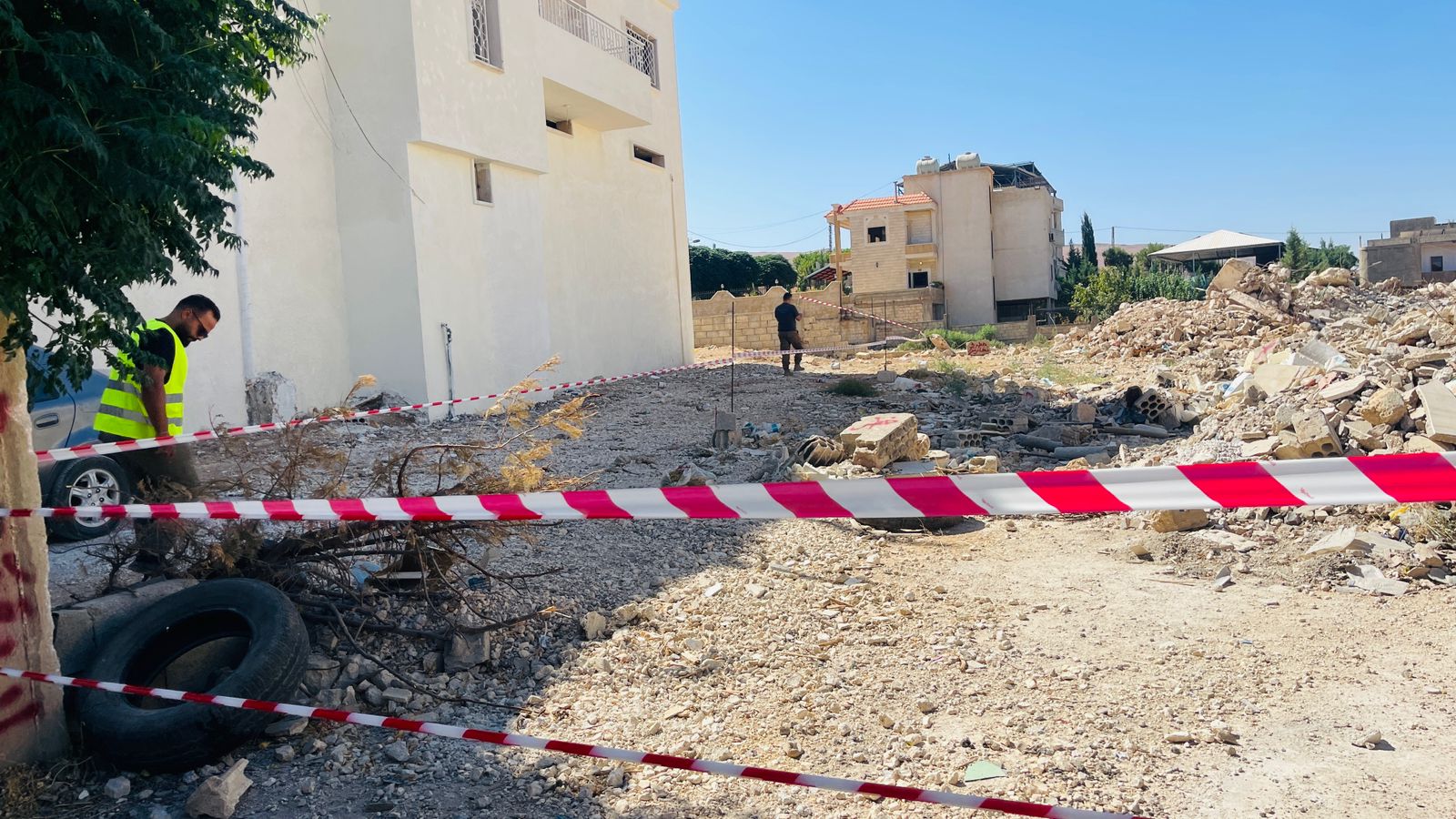 Sur un terrain accidenté au Liban, une rubalise délimite une zone suspectée de contenir des restes explosifs de guerre. Deux personnes s'affairent à l'extérieur de la zone.