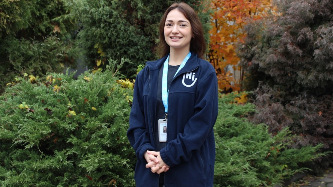 Une femme est debout à l’extérieur, au milieu d’une végétation dense. Elle porte un badge autour du cou, un vêtement bleu foncé avec un logo clair.