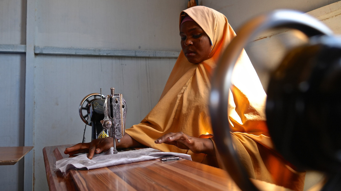 Une jeune femme portant un voile jaune est assise devant une table et coud sur sa machine.