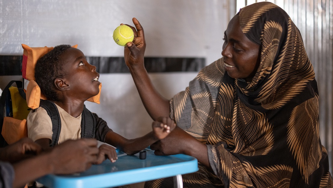 La photo est en intérieur, dans une salle de réadaptation physique. Omran, à gauche, est dans un fauteuil adapté et regarde sa mère Djimilla qui lui montre une balle de tennis. Elle regarde Omran en souriant.
