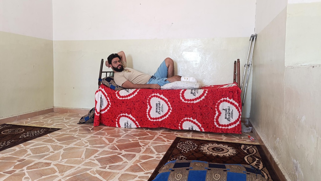 Un jeune homme amputé de la jambe droite est allongé sur un lit et regarde l'objectif. Des béquilles sont posées à côté du lit.