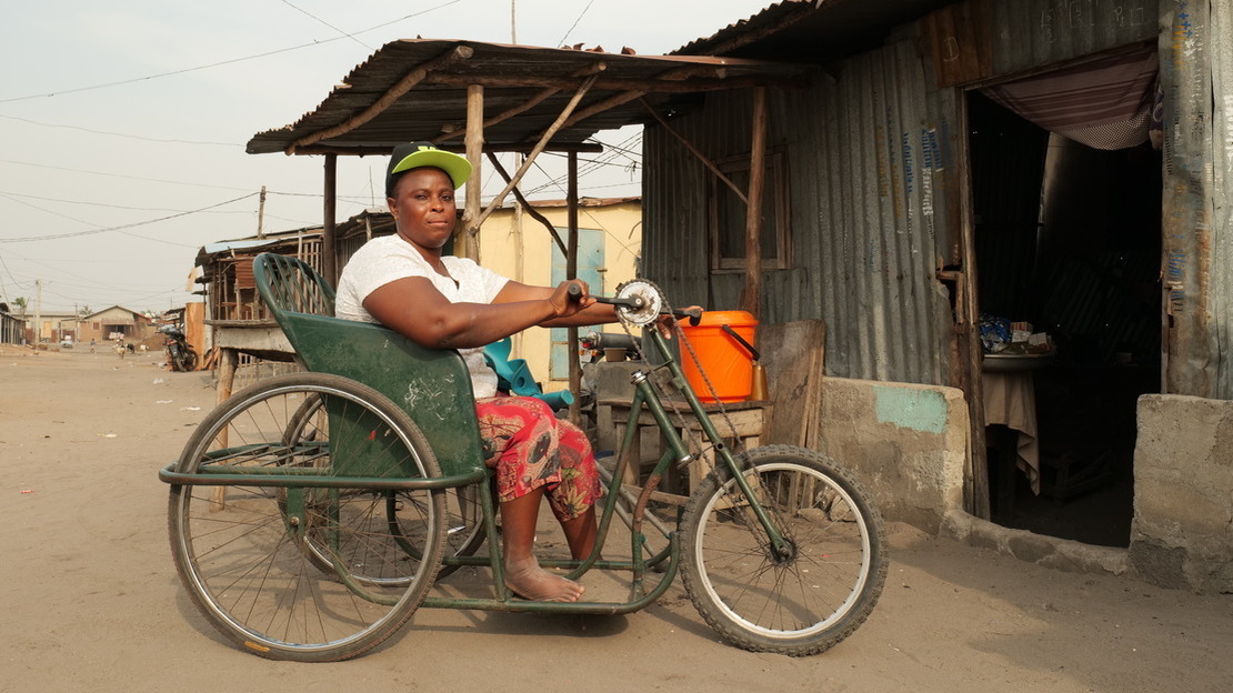 Une femme est assise sur  un tricycle et regarde la photographe. Elle se tient devant un abri fait de tôle et de ciment, et on devine derrière elle une rue et d'autres bâtiments.