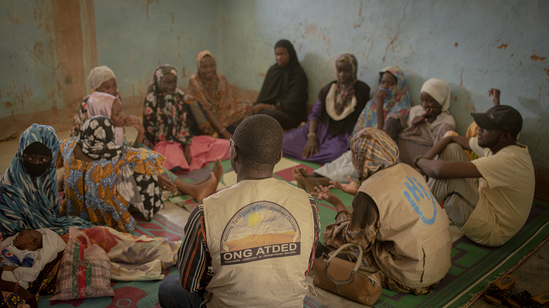 Au Mali, des femmes et des hommes sont assis en cercle sur des nattes, à l'intérieur d'une pièce.