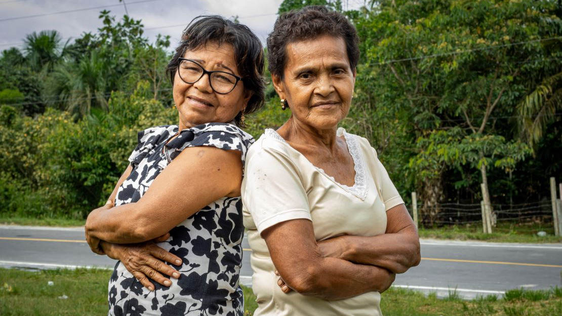 Portrait en gros plan de deux femmes âgées qui regardent le photographe en se faisant une accolade. Derrière elles, une route bitumée et de la végétation.
