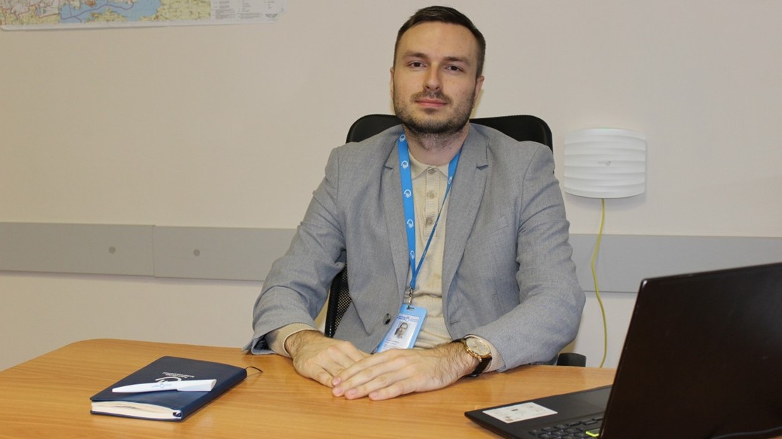Une personne est assise à un bureau . Elle porte une veste claire et un badge avec un cordon bleu. Ses mains sont posées sur le bureau, sur lequel se trouvent un carnet et un ordinateur portable ouvert.