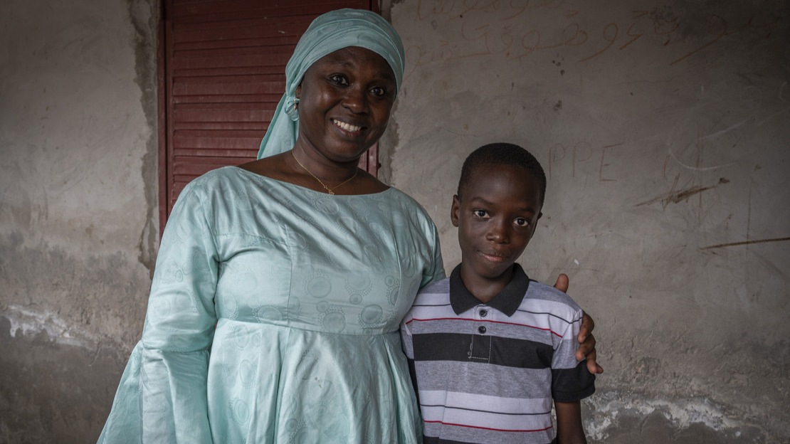 Portrait en gros plan d'une femme qui tient par l'épaule un jeune garçon. Tous deux sourient en regardant le photographe.