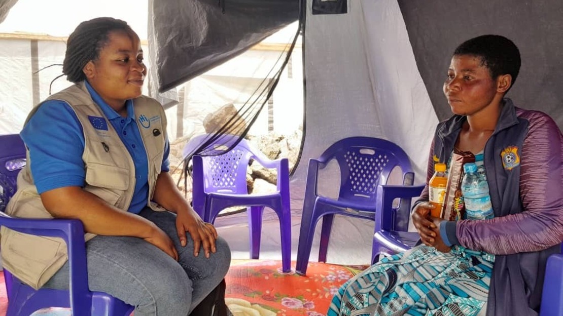 Deux femmes se font face, assises sur des chaises dans une tente. Celle de gauche porte une jaquette floquée HI, celle de droite tient dans ses mains une bouteille d'eau et des biscuits.