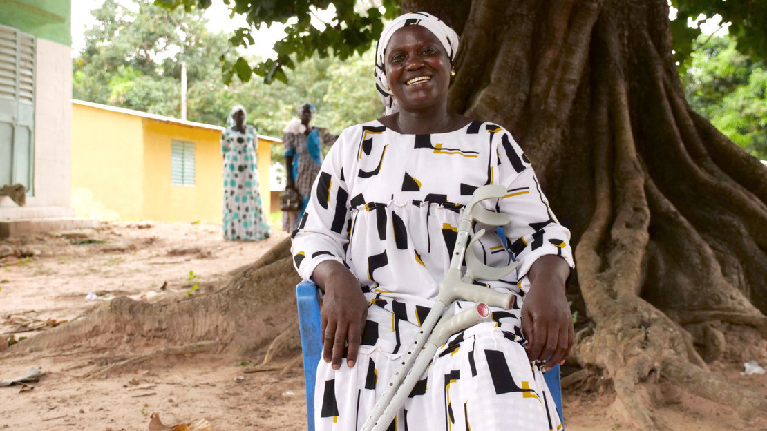 Une femme est assise sur une chaise en plastique bleu, posée devant un grand arbre. Elle est habillée d'une robe blanche aux motifs noir et jaune et sourit. Des béquilles sont appuyées sur ses jambes.