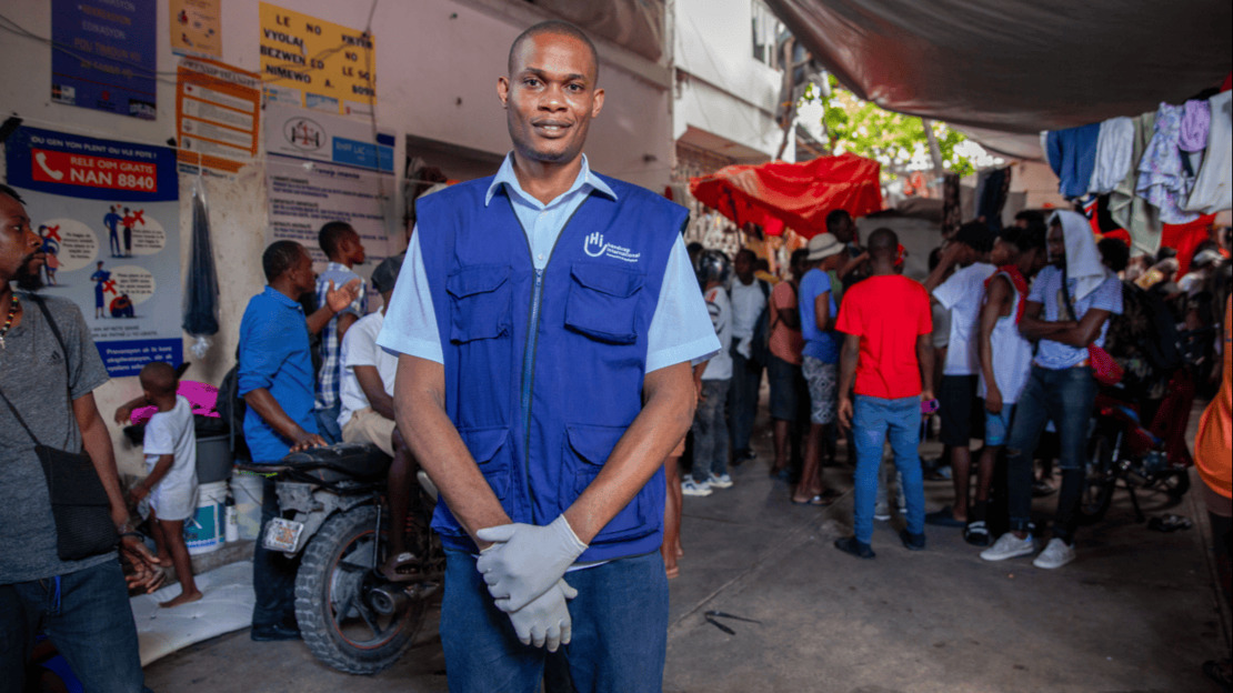 Un homme souriant portant des gants de travail et une jaquette HI. Derrière lui, une salle dans laquelle discutent de nombreuses personnes et où sèche du linge.