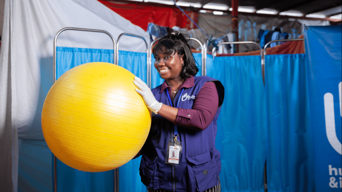 Portrait d'une femme souriante, portant un gant en latex et une jaquette HI, et qui tient à la main un gros ballon jaune de kinésithérapie.