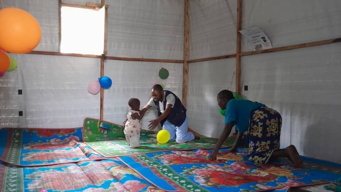 Dans une salle au sol couvert de tapis, un homme ouvre les bras pour tenir un bébé. Une femme accroupie se tient près d'eux. Les murs sont en toile, des ballons y sont accrochés.