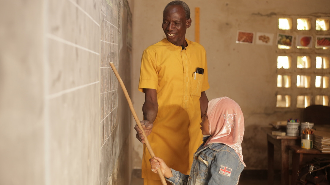 Au Togo, un homme et une fillette sont debout devant un tableau noir dans une classe. Ils tiennent ensemble un bâton qu'ils pointent vers une partie de la leçon écrite au tableau.