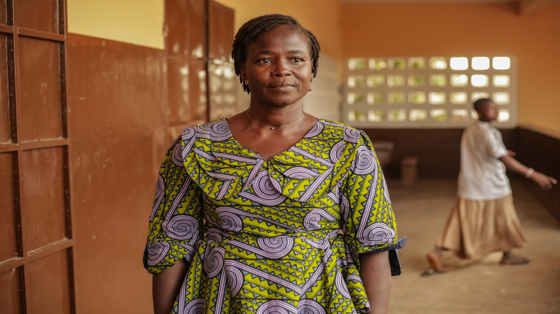 Au Togo, une femme dans un couloir porte une robe à motifs géométriques colorés. Le bâtiment a des murs peints partiellement et des ouvertures laissant entrer la lumière.