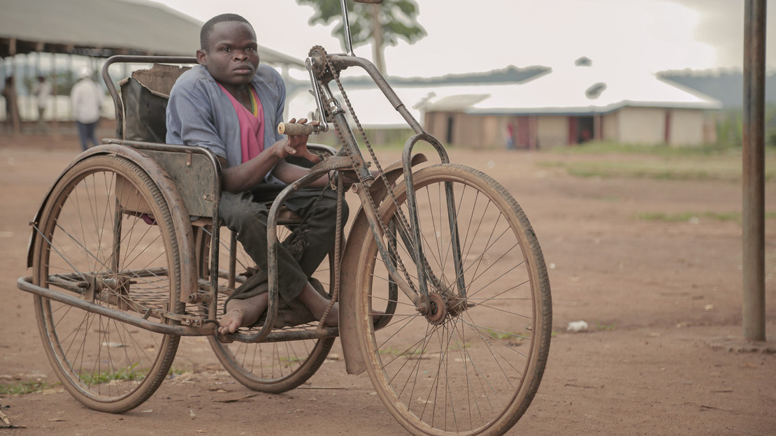 Un homme est assis sur un tricycle et regarde la caméra. Derrière lui, on devine des gens qui marchent, plusieurs bâtiments et un peu de végétation.