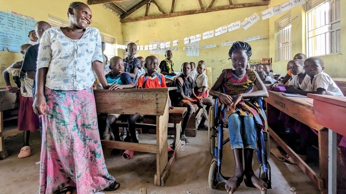 Vue d'une salle de classe avec des élèves assis à des pupitres. Au premier plan, une femme adulte sourit en regardant à sa gauche une jeune fille assise sur un fauteuil roulant qui sourit également.