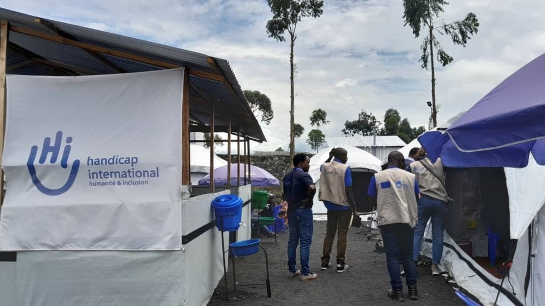 Quatre personnes sont de dos dans une ruelle bordée de deux tentes. Sur celle de gauche est accroché un large drapeau aux couleurs de Handicap International. Au second plan on voit d'autres tentes.