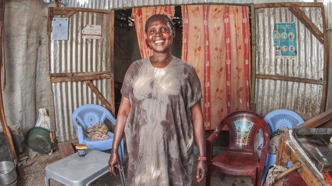 Une femme se tient debout dans une pièce aux murs de tôle et de bois, entourée de chaises et de tables en plastique. Elle a un large sourire et regarde le photographe.