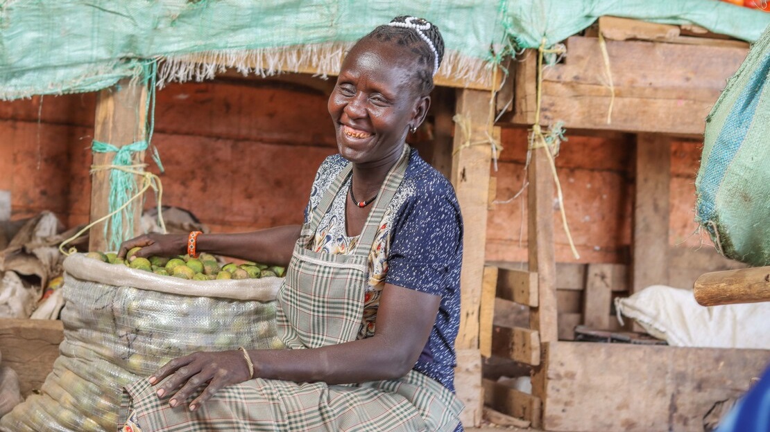 Une femme est assise et rigole. Elle a la main droite posée sur un grand sac en toile contenant des fruits. Derrière elle, on devine des étales de fruits et légumes.