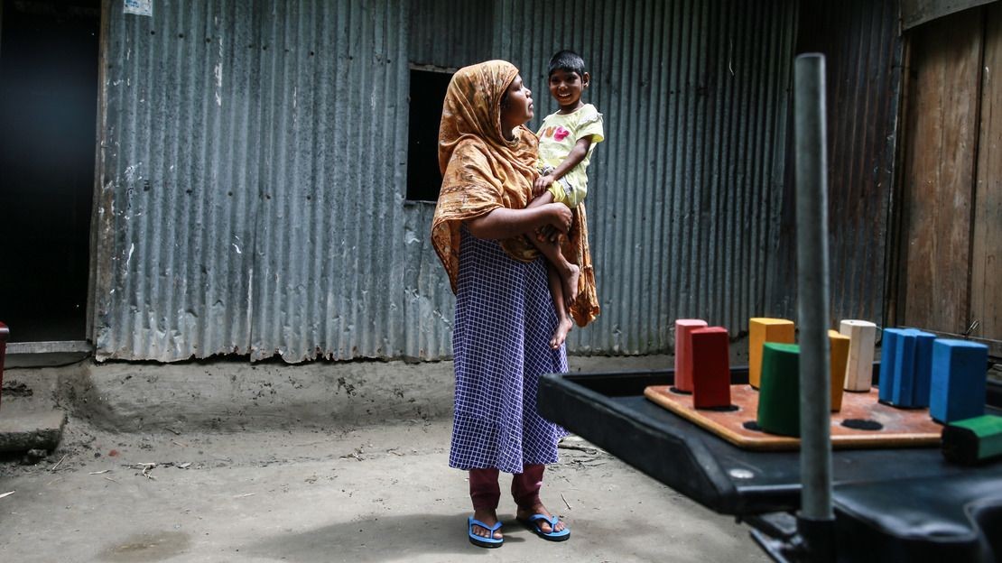 Dans une maison de tôles, une mère porte sa fille handicapée qui sourit. Au premier plan, un jouet thérapeutique en bois de plusieurs objets de couleur.