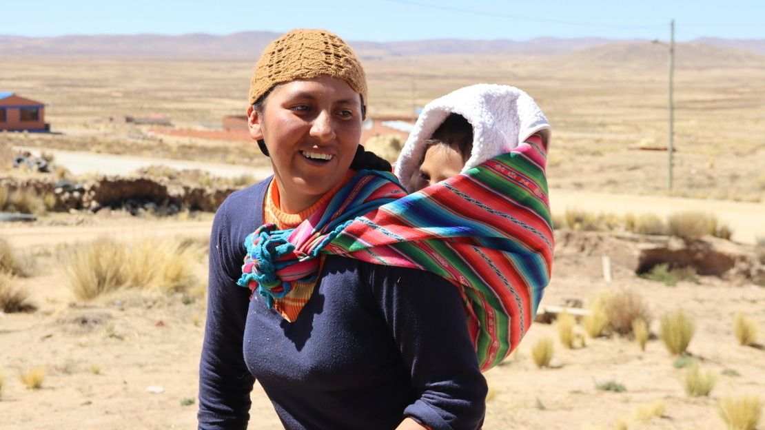 Une femme souriante porte son fils dans son dos à l'aide d'un tissu traditionnel (aguayo). En arrière-plan, des plaines sèches et poussiéreuses.