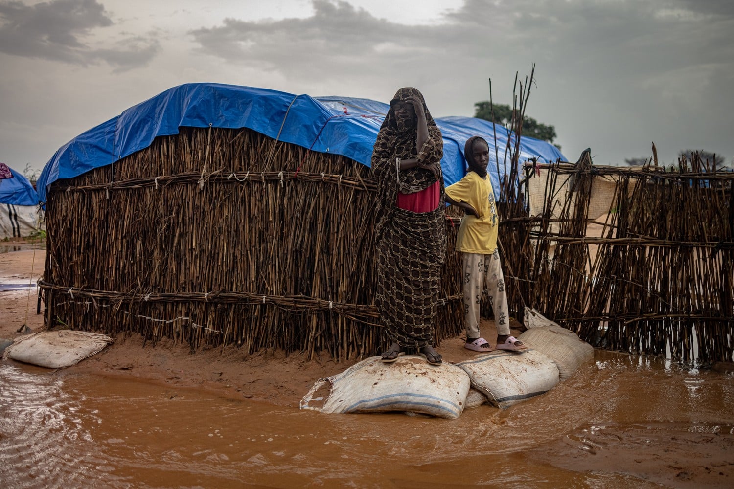 Entourées d'eau et de boues, une femme et une jeune fille se tiennent debout sur des sacs devant une maison de bois dont le toit est recouvert d'une bâche bleue.