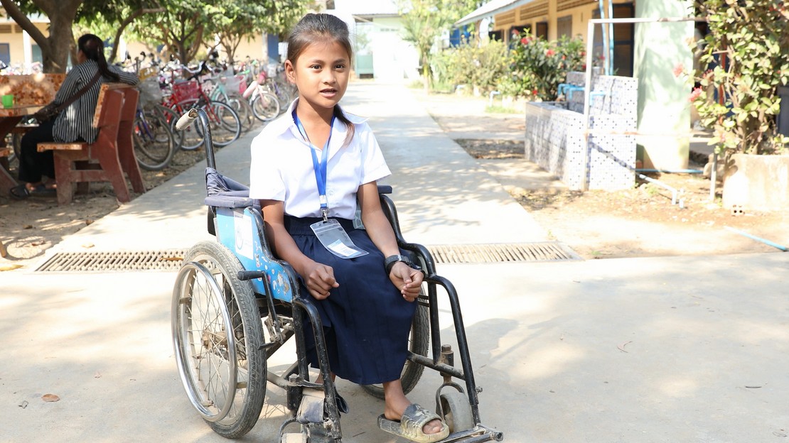 Une jeune fille est assise dans un fauteuil roulant. Elle porte une chemise blanche à manches courtes, un pantalon sombre et un badge autour du cou.  À l’arrière-plan, plusieurs vélos sont stationnés en rangée le long du chemin.