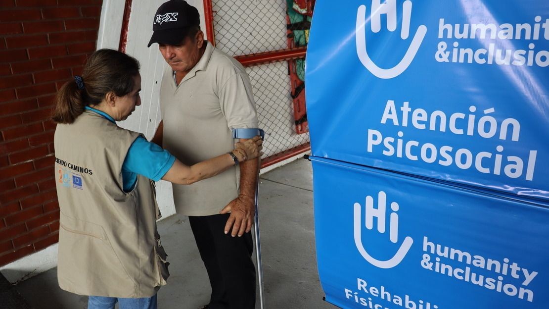 Une femme de dos aide un homme à tenir une béquille.