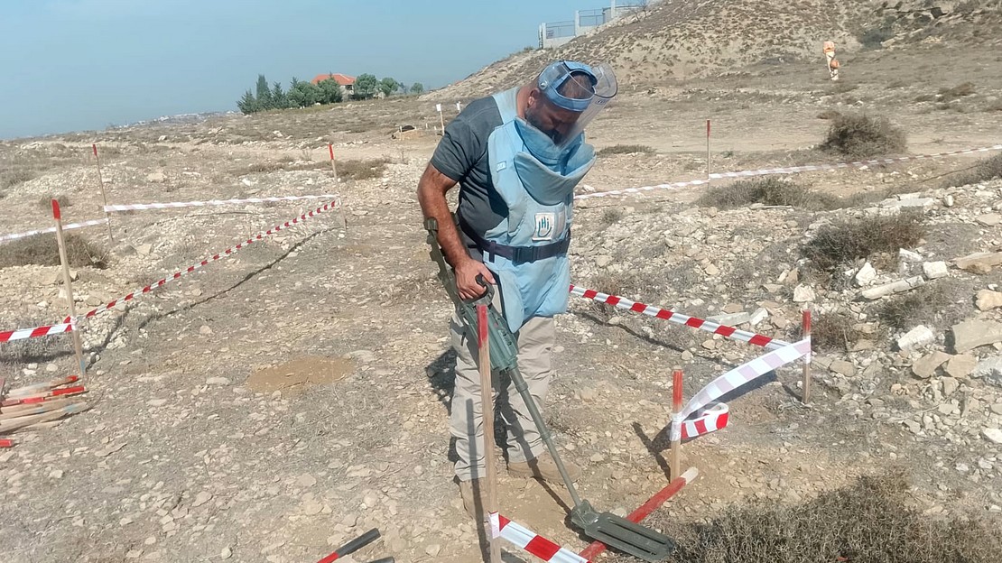 Un démineur en tenue de protection se tient debout sur un terrain aride et escarpé. Il manipule de sa main droite un détecteur de métaux et prend garde à rester dans la zone sécurisée, délimitée par du rubalise rouge et blanc.