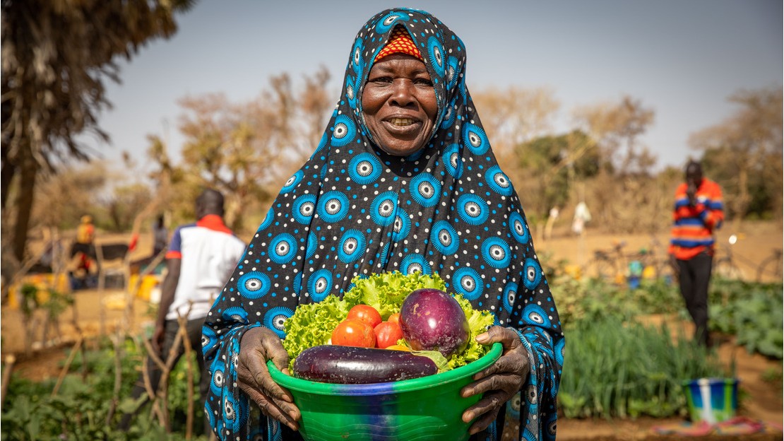 Fadima pose avec dans la main une bassine qui contient des tomates, des aubergines et de la salade. En arrière-plan, on devine le jardin partagé.