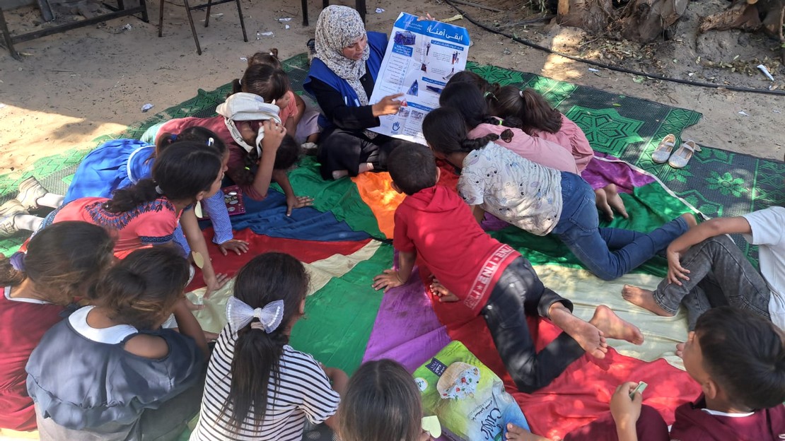 Dans un camp de déplacés à Gaza, une femme tient une affiche et montre à plusieurs enfants très attentifs les dangers des restes explosifs de guerre.