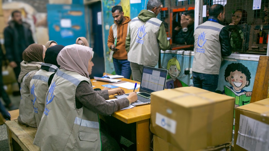 Dans une pièce, plusieurs personnes avec un gilet HI organisent la distribution de cartons contenant des kits d'hygiène pour des réfugiés fuyant les combats.