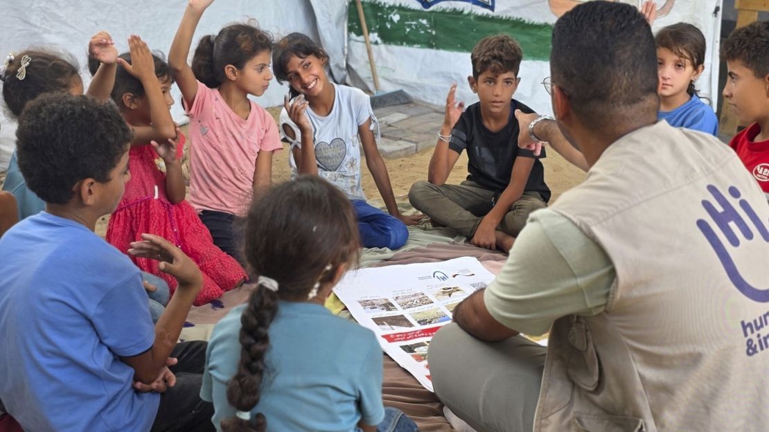 Plusieurs enfants sont assis par terre autour d'une affiche de sensibilisation aux dangers des restes explosifs de guerre. De dos, un homme les interroge et leur donne la parole un par un.