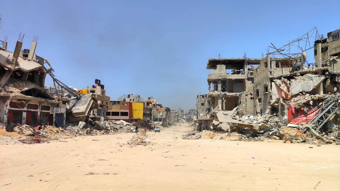 Bâtiments presqu'entièrement détruits de part et d'autre d'une voie de circulation recouverte de sable. Au loin on devine un véhicule et quelques piétons.
