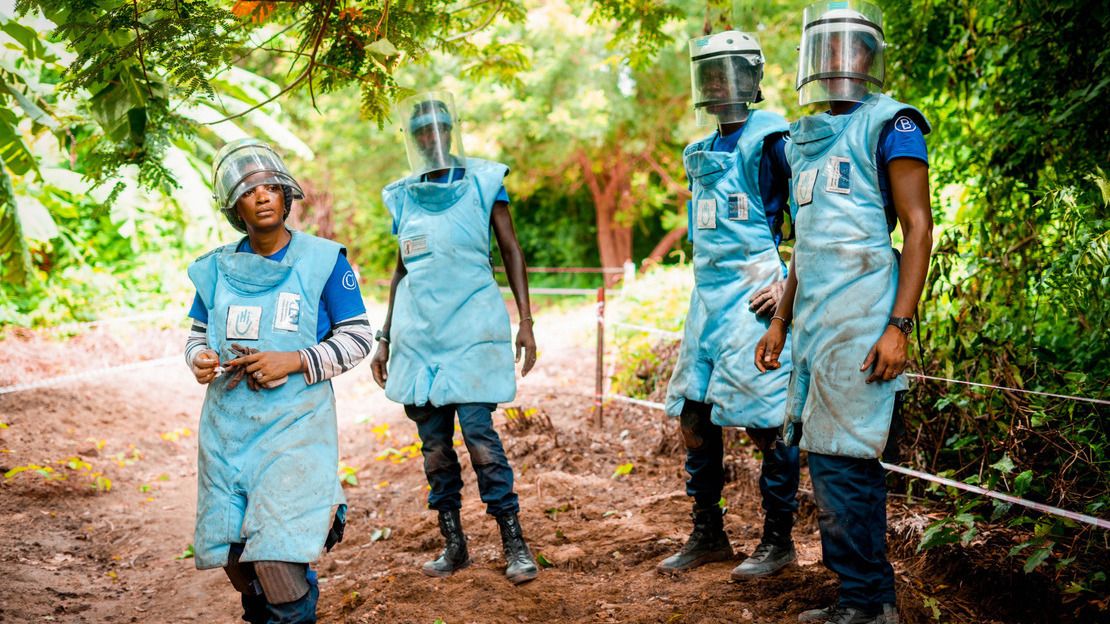 Trois démineurs et une démineuse en tenue de protection se tiennent sur un chemin de terre, entourés de végétation luxuriante.