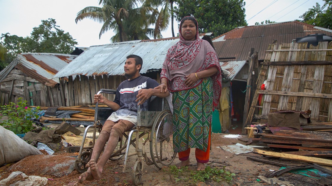 Devant des maisons touchées par des inondations au Bangladesh, un homme en fauteuil roulant et une femme à ses côtés.
