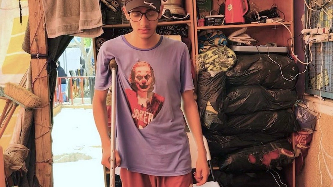 Dans un abri temporaire, un jeune homme portant une casquette et des lunettes se tient debout appuyé sur une béquille.