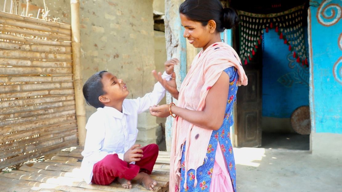Un petit garçon est assis et tend le bras vers sa mère habillée en tenue traditionnelle qui lui sourit devant leur maison au Népal.