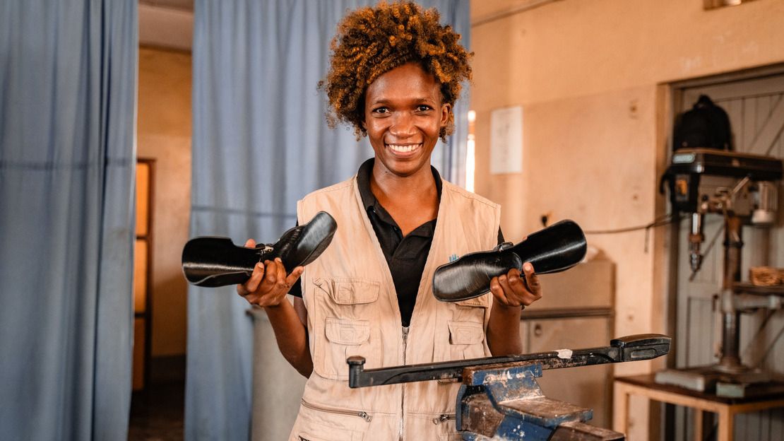 Une jeune femme noire aux cheveux bouclés est dans un atelier d'appareillage. Elle tient dans chacune de ses mains une orthèse en plastique noir et sourit.