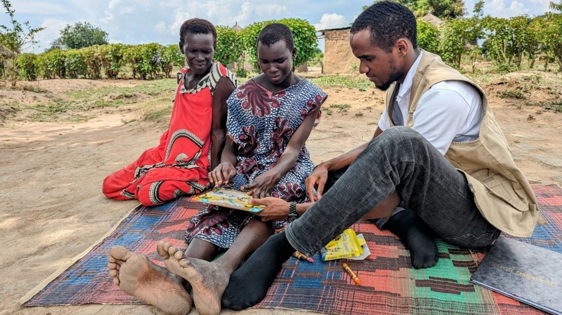 Santina faisant un puzzle, accompagnée de sa mère et d'un ergothérapeute de Handicap International. Ils sont assis tous les trois sur un tapis, à l'extérieur. Santina est au centre de l'image, et sa mère et l'ergothérapeute, qui l'entourent,  la regardent en faire le puzzle en souriant.