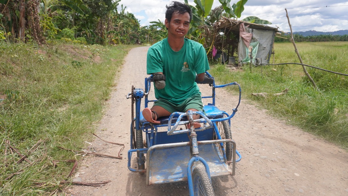 Aux Philippines, un jeune homme amputé des quatre membres est assis sur un tricycle sur un chemin de terre.