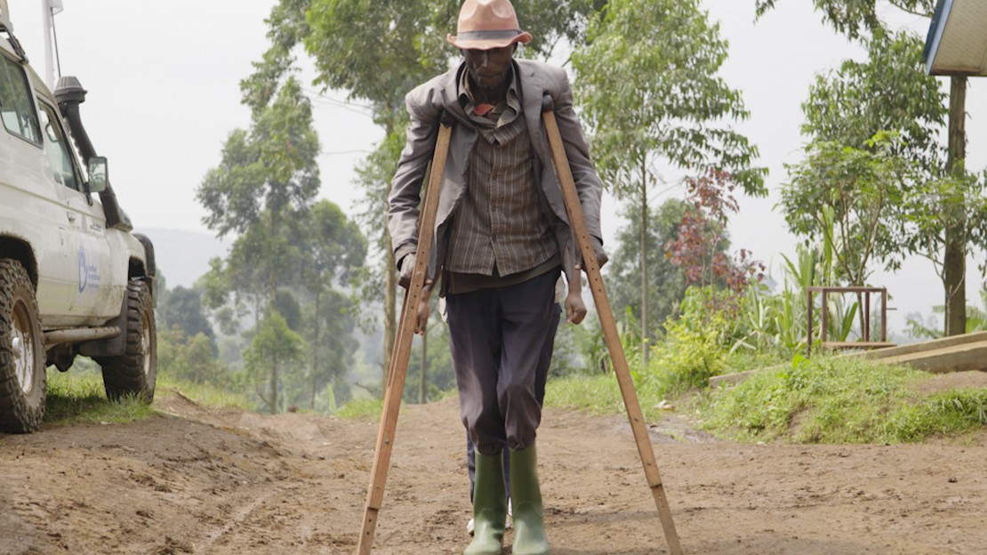 En République démocratique du Congo, un homme âgé avance sur un chemin en terre en s’appuyant sur deux longues béquilles en bois, placées de chaque côté de son corps.