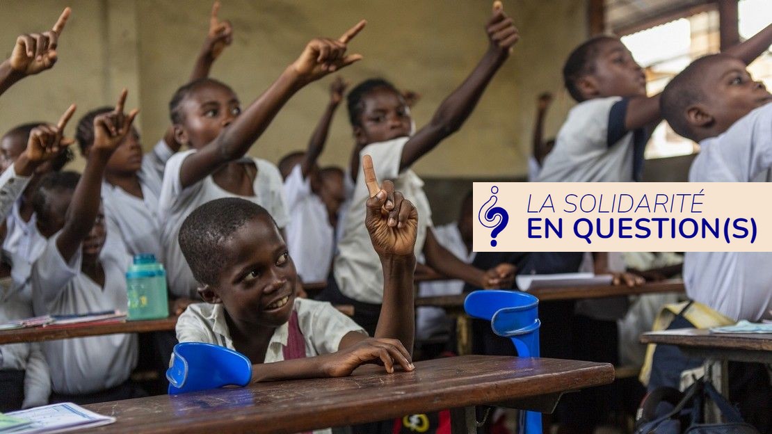 Dans une salle de classe en République démocratique du Congo. Au premier plan, un enfant est assis et lève le doigt ; à côté de lui sont posées ses béquilles bleues. Au second plan, d'autres élèves lèvent également le doigt, certains sont assis et d'autres sont debout.