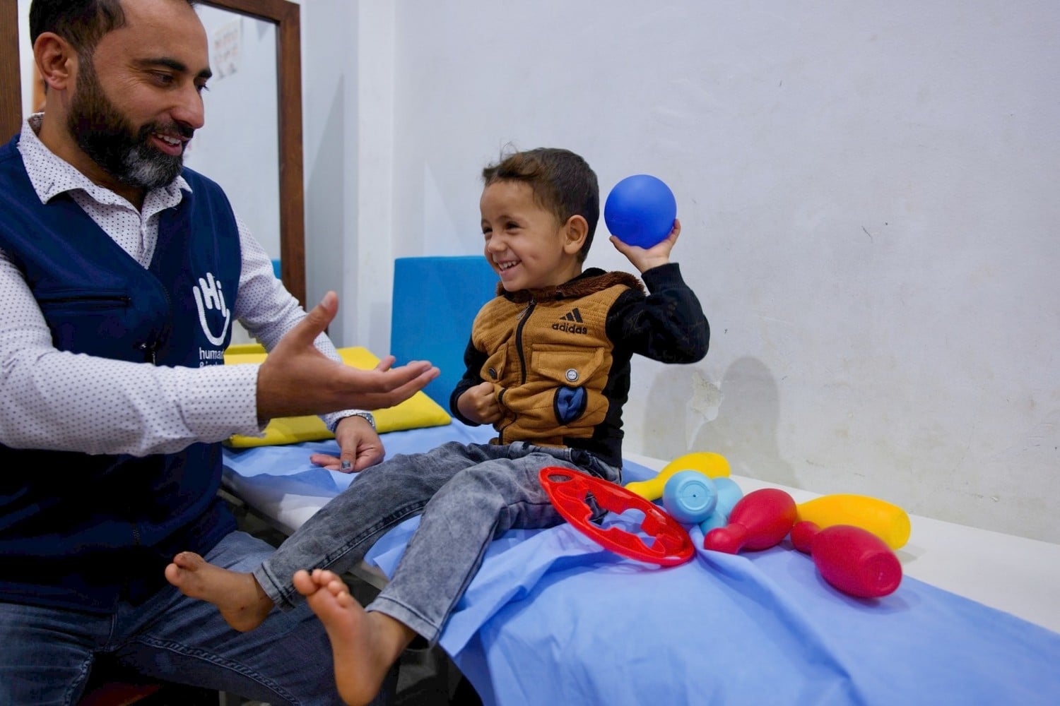 Assis sur une table d'auscultation, un jeune garçon souriant joue avec une balle et s'apprête à la lancer à un homme assis à côté de lui.