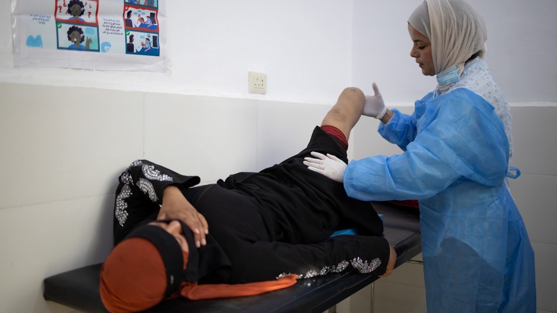 En Syrie, une jeune femme est allongée sur une table d’examen dans une pièce médicale. Elle est amputée d’une jambe et le moignon est visible. À côté de la table, une autre personne porte une tenue médicale et manipule le moignon avec ses mains.