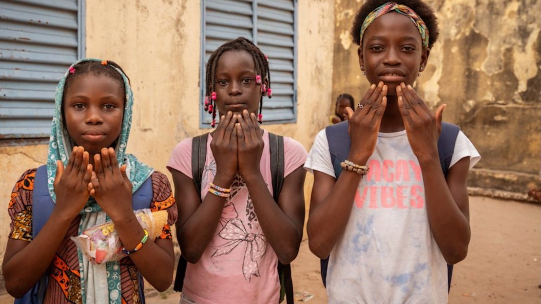 Trois jeunes filles se tiennent debout côte à côte et portent chacune leurs deux mains au niveau du bas de leur visage, paumes ouvertes tournées vers elles.