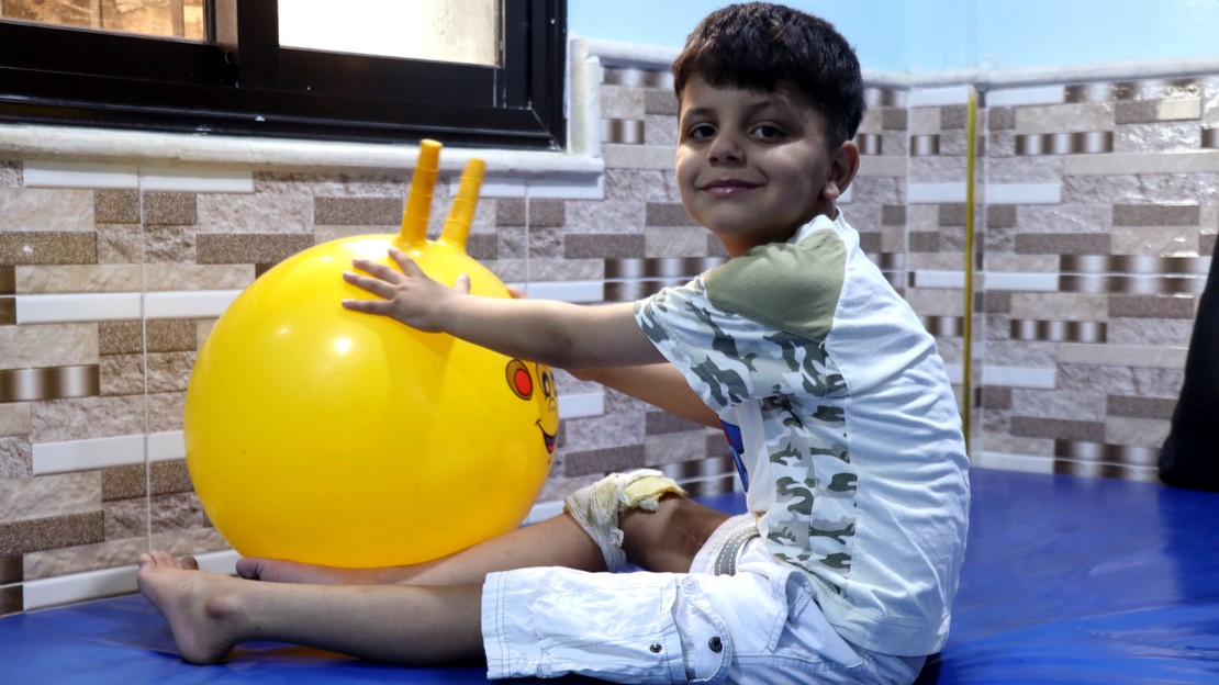 Un jeune garçon est assis sur un tapis et utilise un ballon pour l'aider dans sa rééducation.