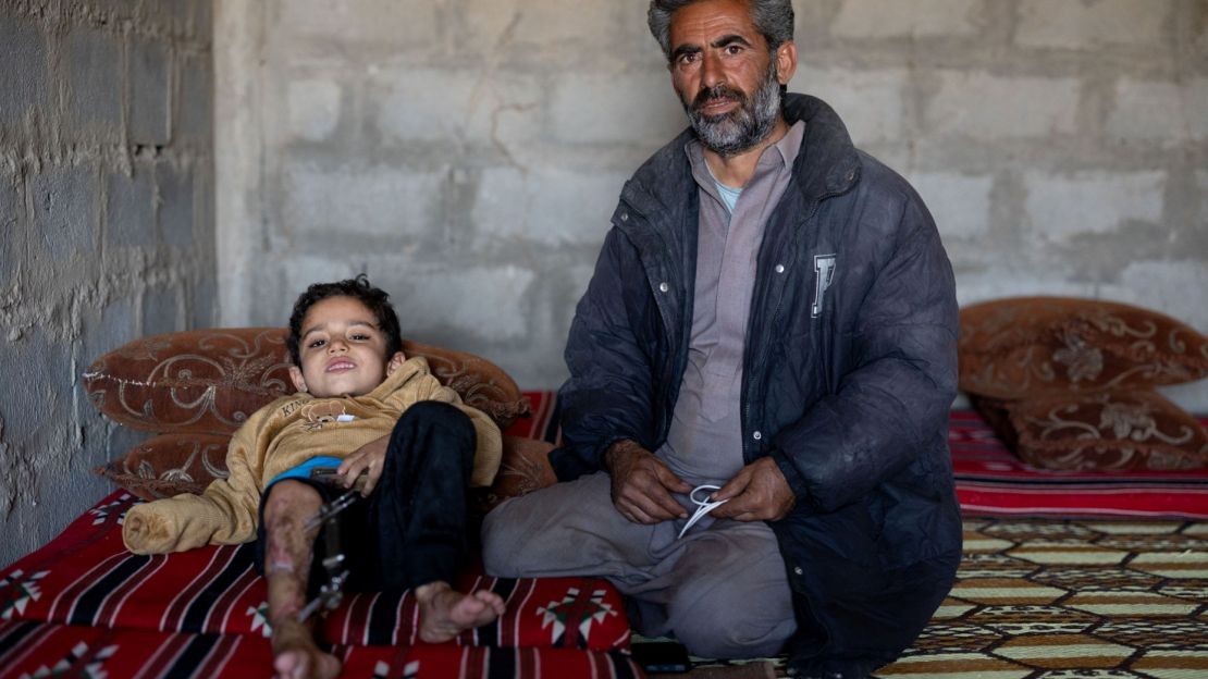 Deux personnes se trouvent à l’intérieur d’une pièce. Un homme est assis au sol sur un tapis, légèrement tourné vers le photographe, avec un objet blanc tenu entre les mains. À côté, un enfant est allongé sur un matelas . L’une de ses jambes est équipée d’un dispositif médical et l’autre jambe présente des blessures.